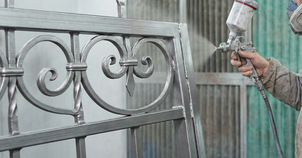 専用の塗装ブース内で、防塵マスクを着用した作業員がスプレーガンを使い、下地処理を終えたアイアンゲートに銀色の防錆塗料を均一に吹き付けている様子。